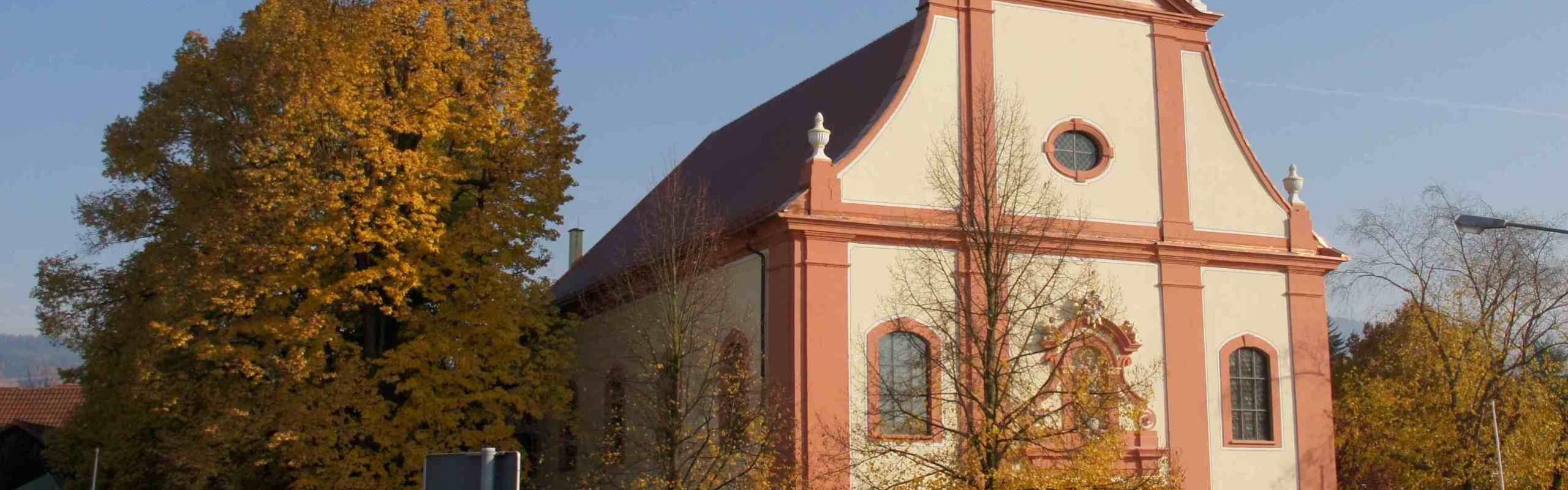 Das Bild zeigt eine Außenansicht einer Kirche mit einem herbstlichen großen Baum n